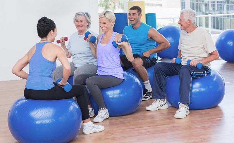 Gruppe von Kursteilnehmern sitzt auf Gymnastikbällen und trainiert mit Physiotherapeutin